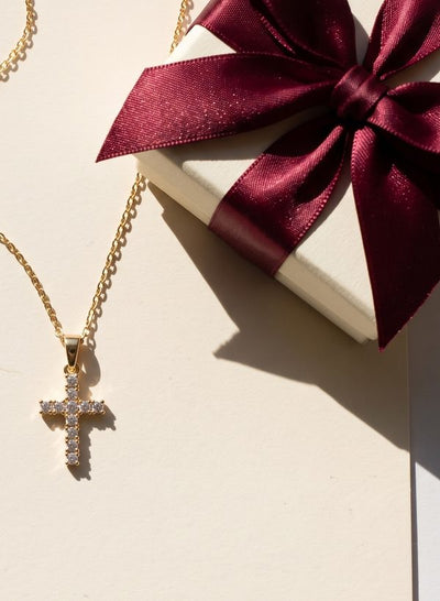 Gold cross necklace with clear stones lying on an ivory card, beside an ivory gift box tied with a burgundy satin bow