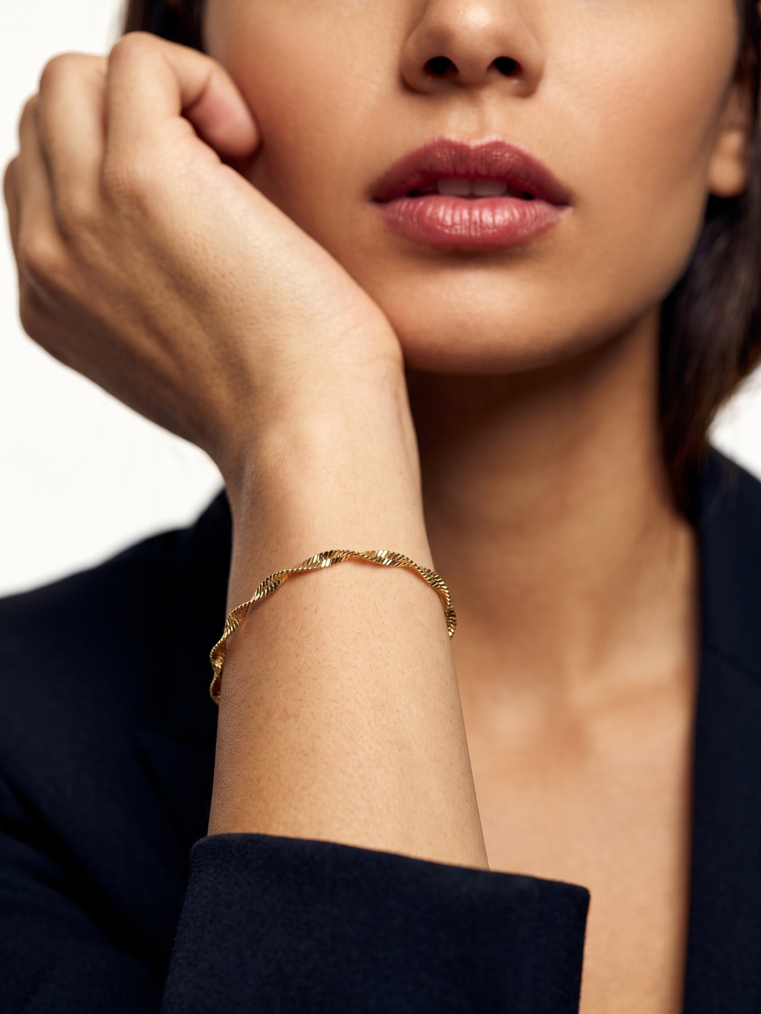 Anchor Twist Chain Bracelet – worn on the wrist, hand raised to the cheek, gold, dark blazer, ivory studio background