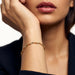 Anchor Twist Chain Bracelet – worn on the wrist, hand raised to the cheek, gold, dark blazer, ivory studio background