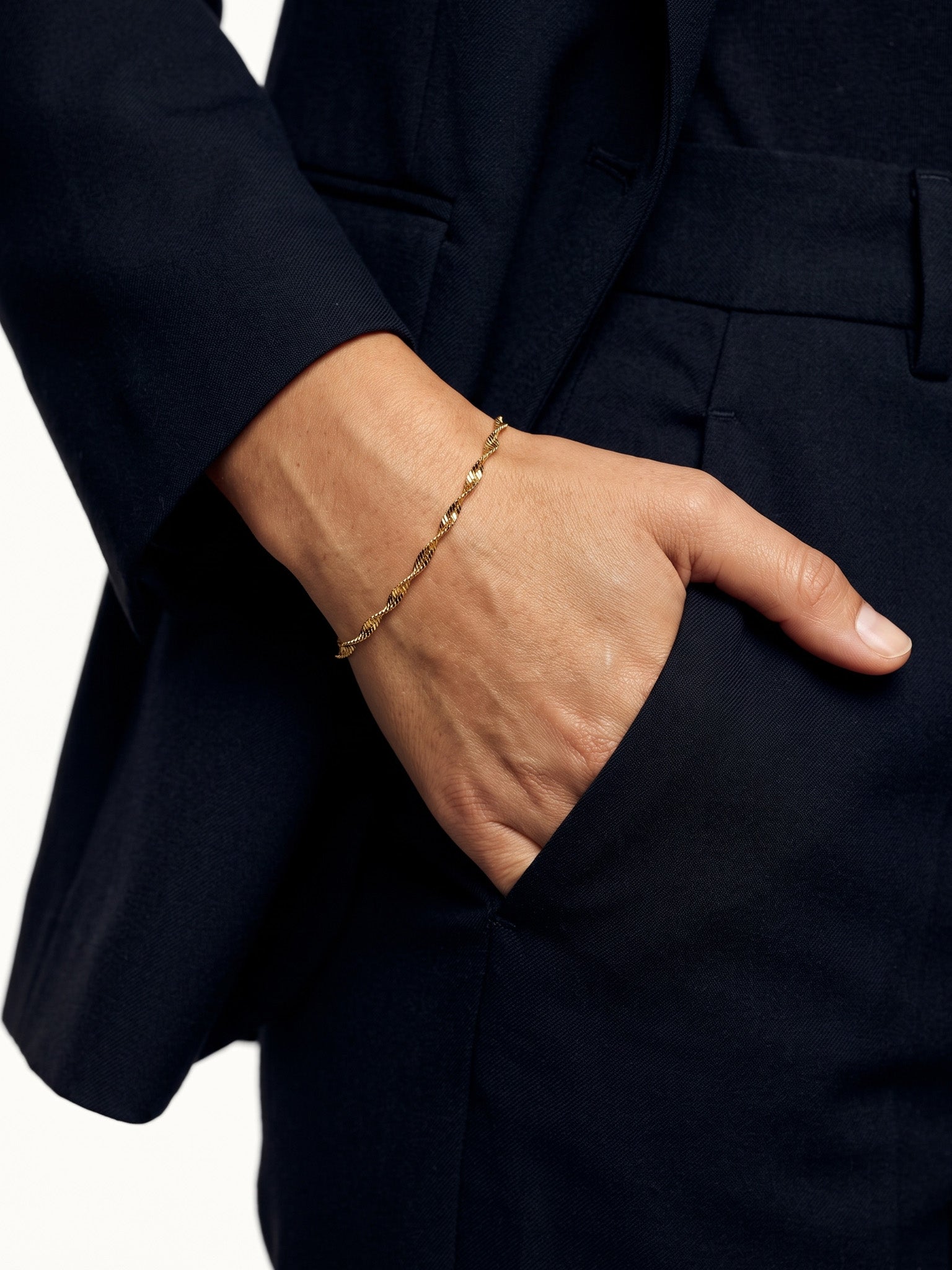 Anchor Twist Chain Bracelet – worn on the wrist, hand in the trouser pocket, gold, dark blazer, ivory studio background