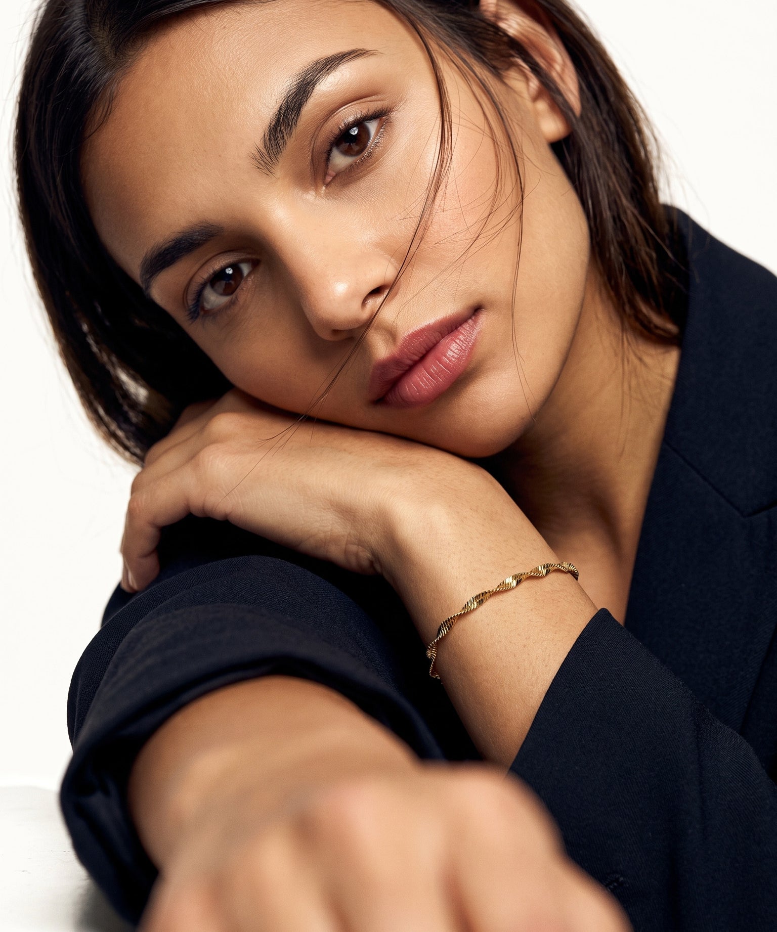 Anchor Twist Chain Bracelet – worn on the wrist, head resting on the arm at a table, gold, dark blazer, ivory studio background