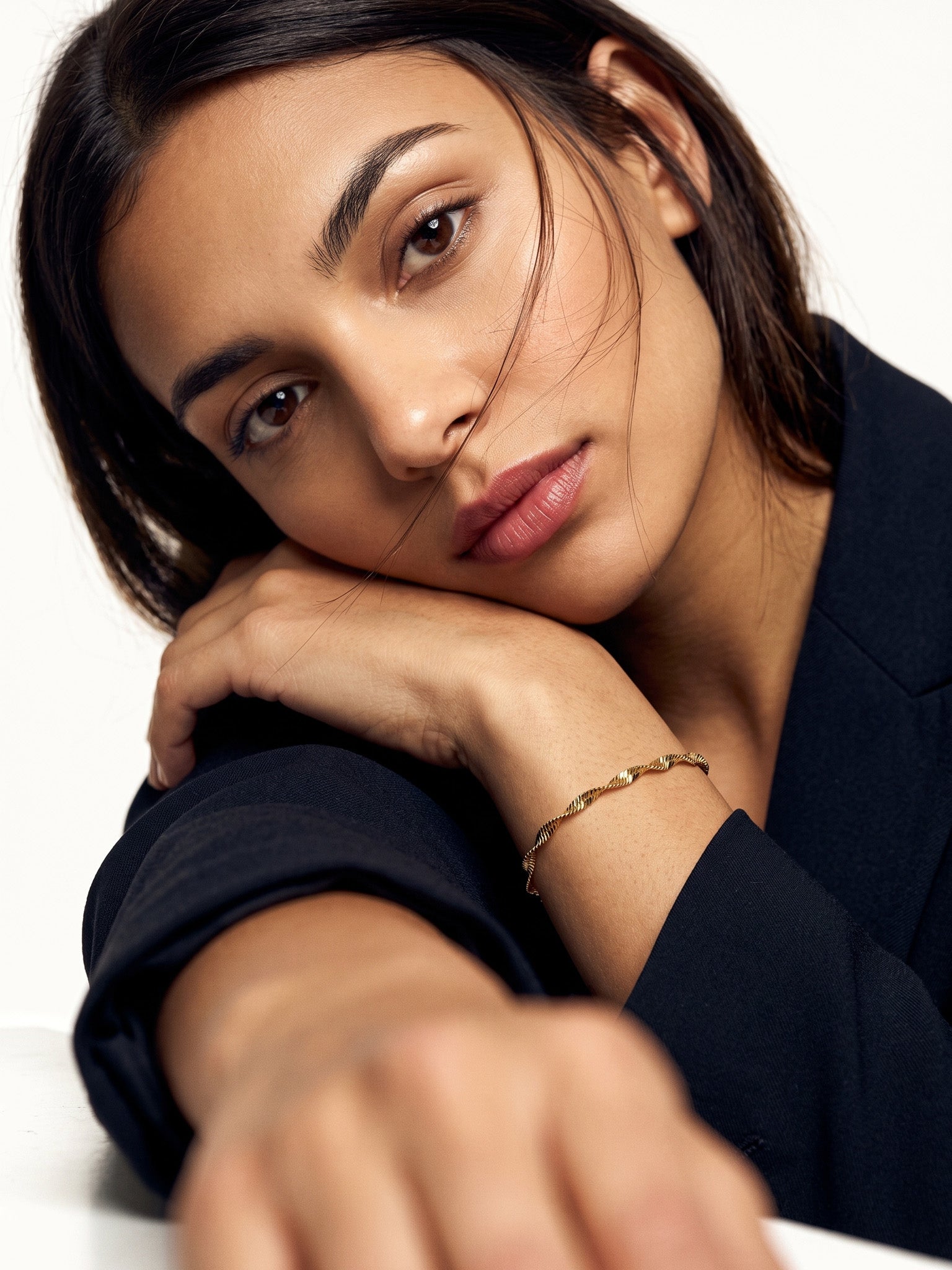 Anchor Twist Chain Bracelet – worn on the wrist, head resting on the arm at a table, gold, dark blazer, ivory studio background