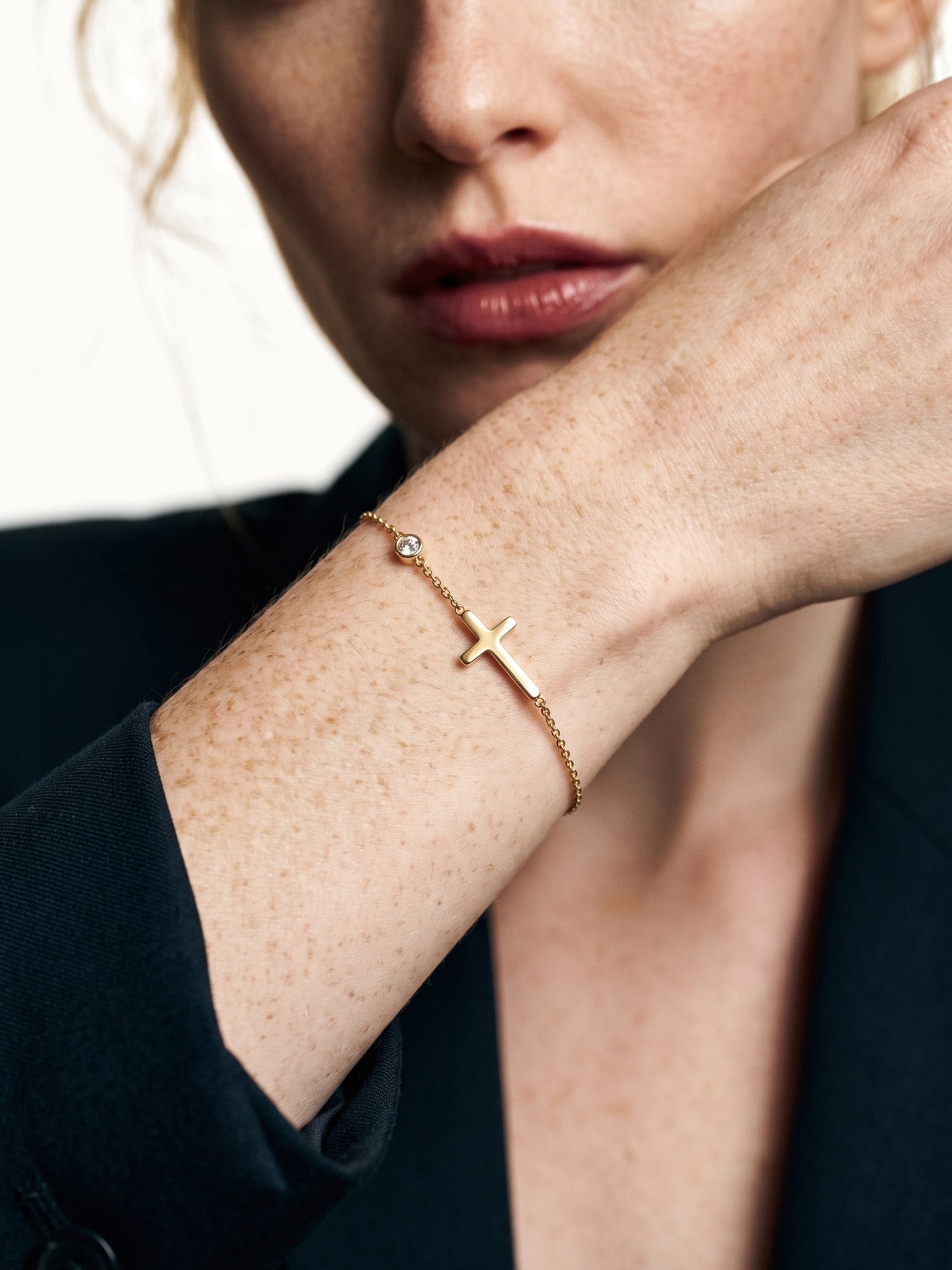 Beside Me Cross Bracelet in gold, worn on the wrist, close-up with the hand raised to the face, dark blazer, ivory studio background