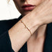 Beside Me Cross Bracelet in gold, worn on the wrist, close-up with the hand raised to the face, dark blazer, ivory studio background