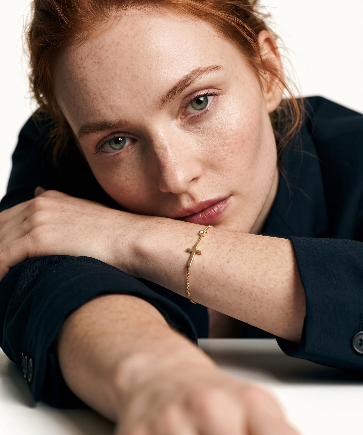 Beside Me Cross Bracelet in gold, worn on the wrist, head resting on the arm at a table, dark blazer, ivory studio background