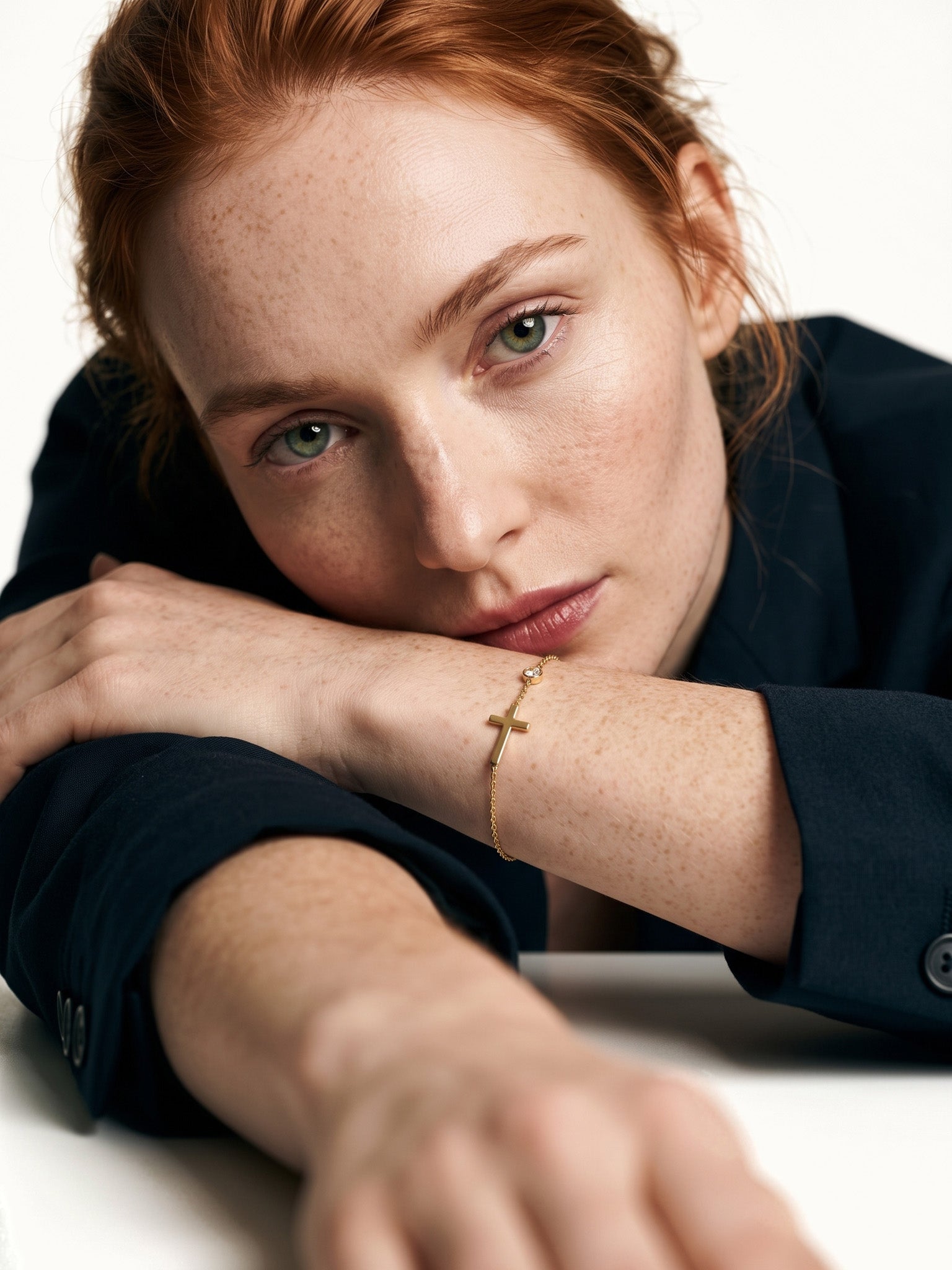 Beside Me Cross Bracelet in gold, worn on the wrist, head resting on the arm at a table, dark blazer, ivory studio background