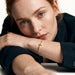 Beside Me Cross Bracelet in gold, worn on the wrist, head resting on the arm at a table, dark blazer, ivory studio background