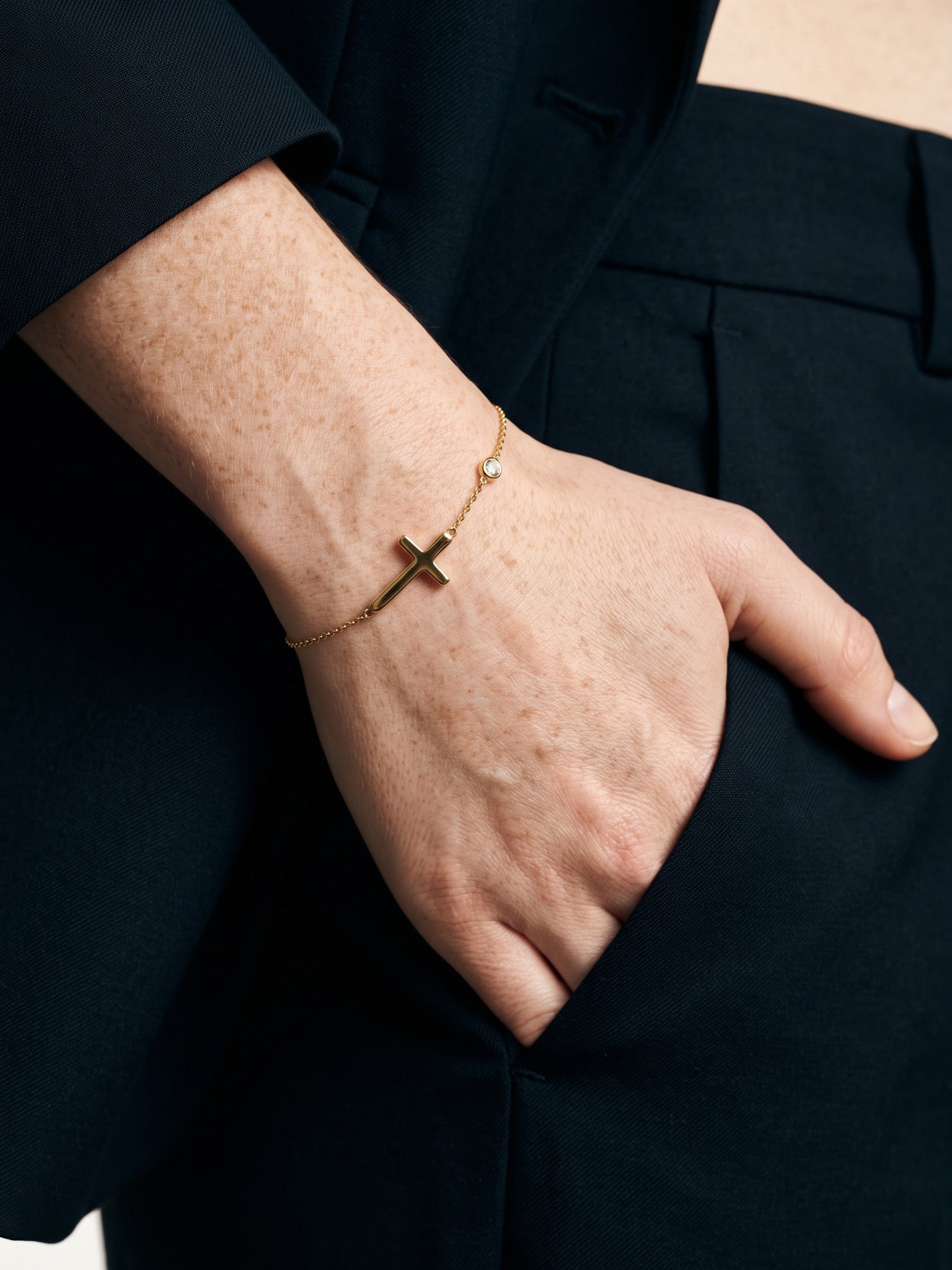 Beside Me Cross Bracelet in gold, worn on the wrist, hand in the trouser pocket, dark blazer, ivory studio background