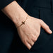 Beside Me Cross Bracelet in gold, worn on the wrist, hand in the trouser pocket, dark blazer, ivory studio background
