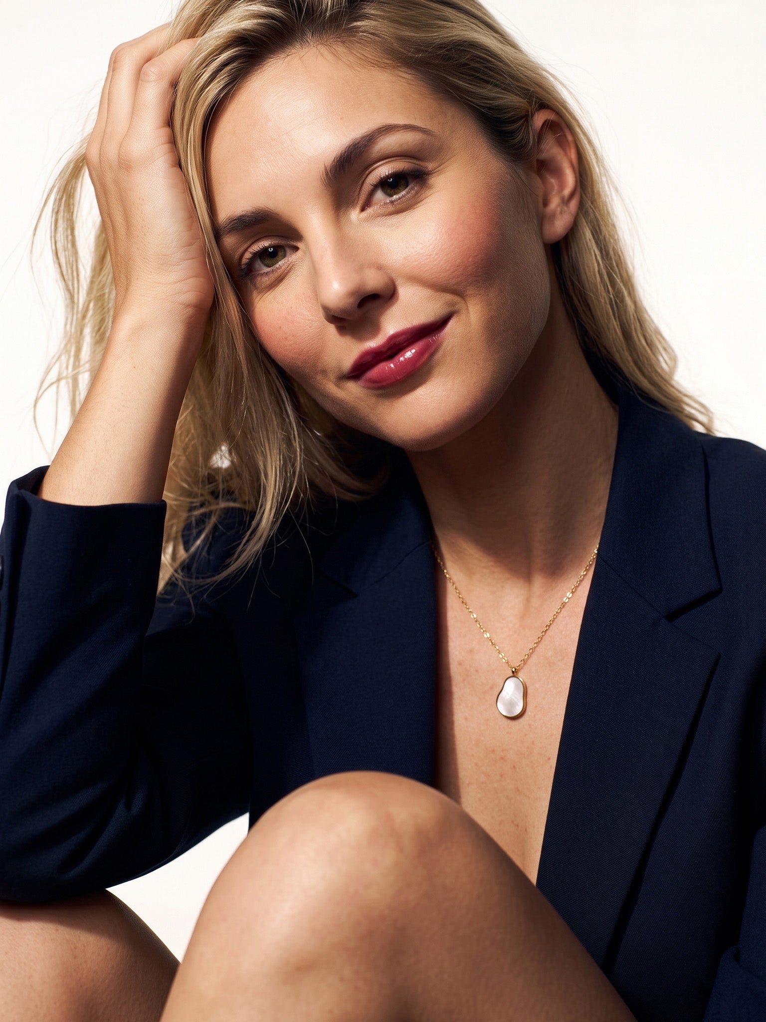 Dawn Pearl Pendant Necklace – worn on the chest, seated with a hand in her hair, navy blazer, mother-of-pearl pendant, ivory studio background