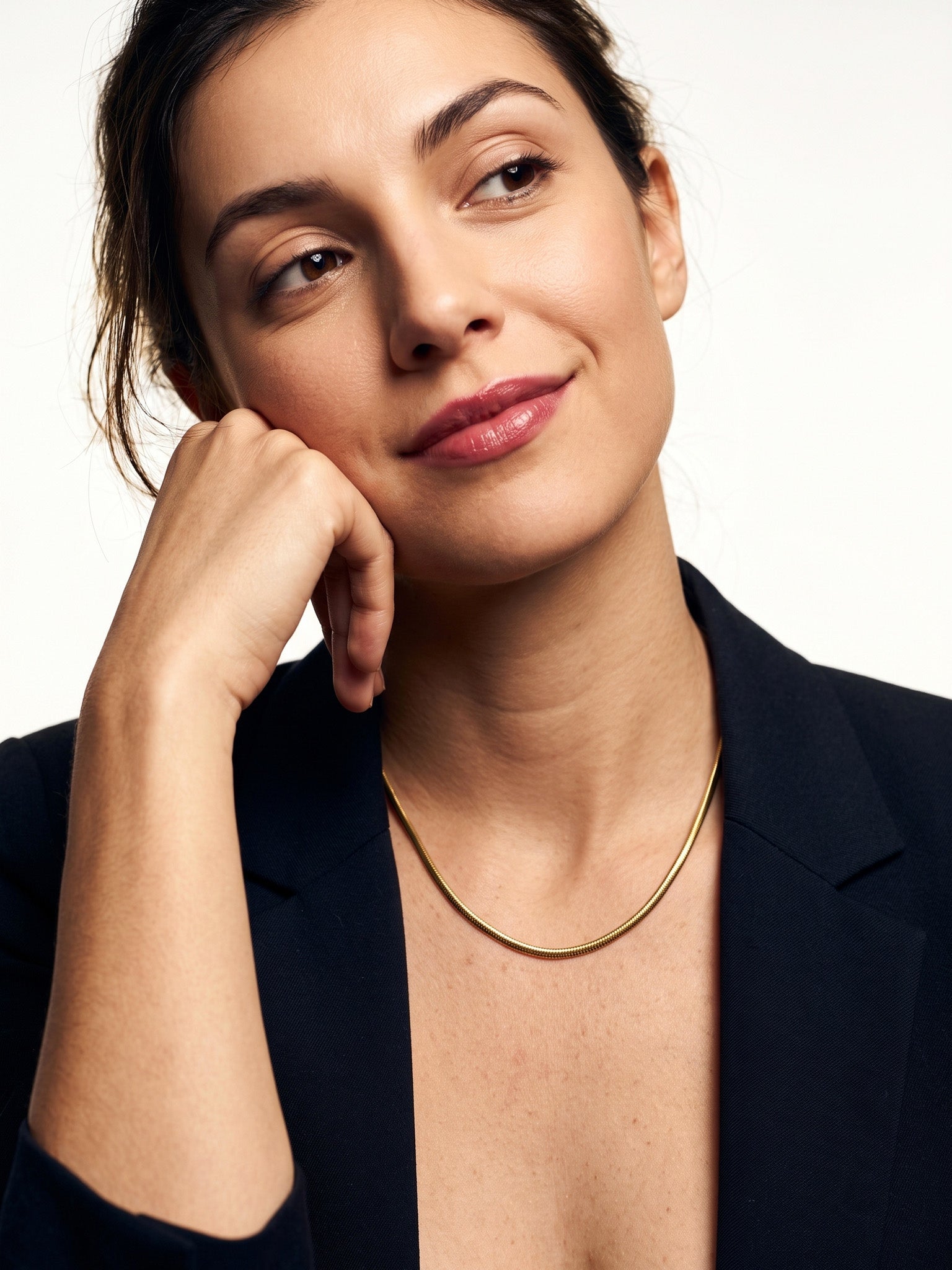 Everlasting Chain Necklace – worn at the collarbone, head resting on a fist, gold, black blazer, ivory studio background