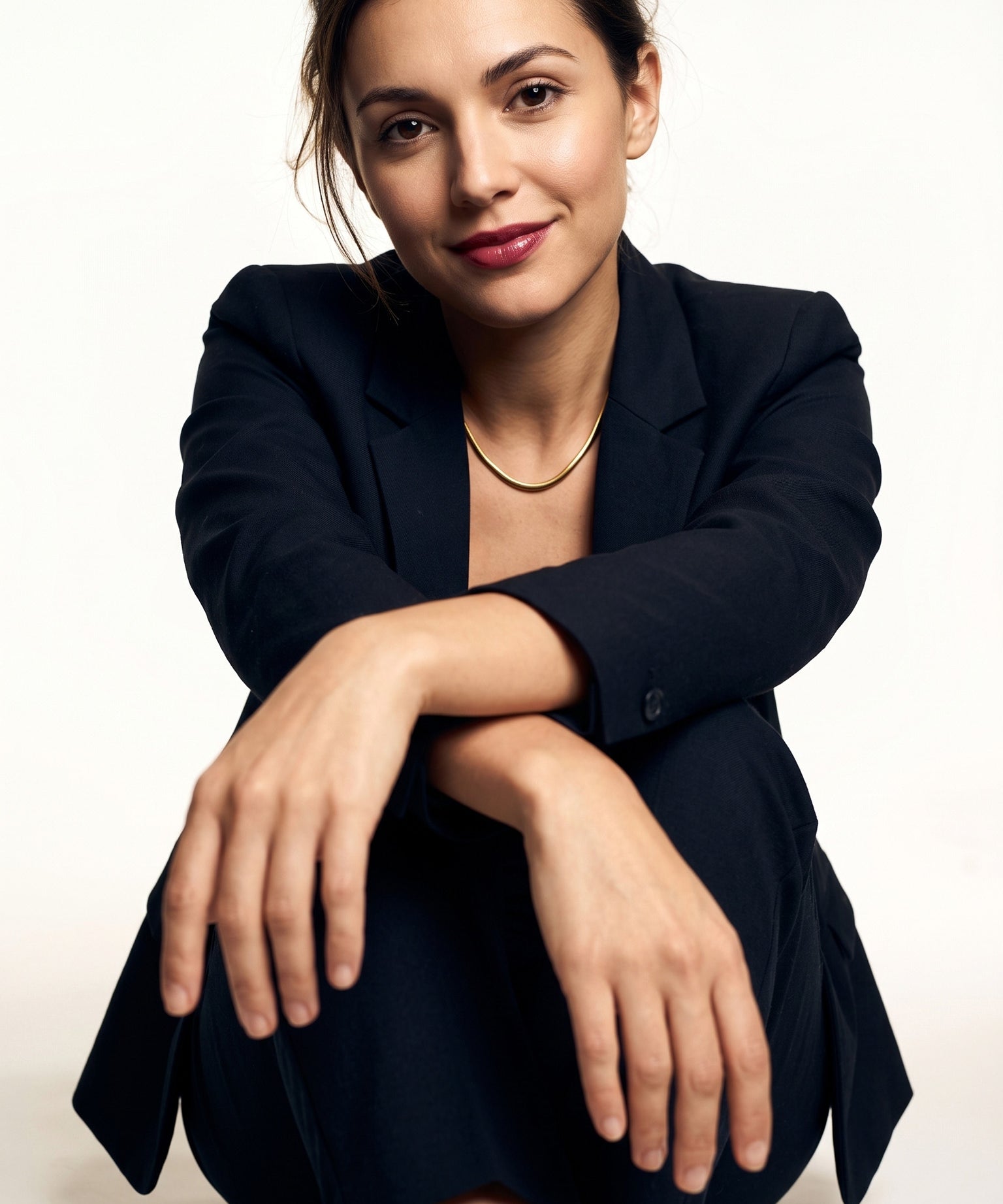 Everlasting Chain Necklace – worn at the collarbone, seated with an arm across the knee, gold, black suit, ivory studio background