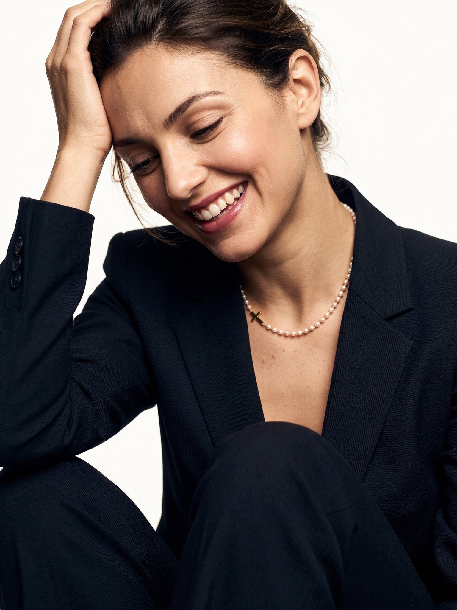 Every Season Cross Necklace – worn on the neck, laughing with one hand in the hair, gold, dark blazer, ivory studio background