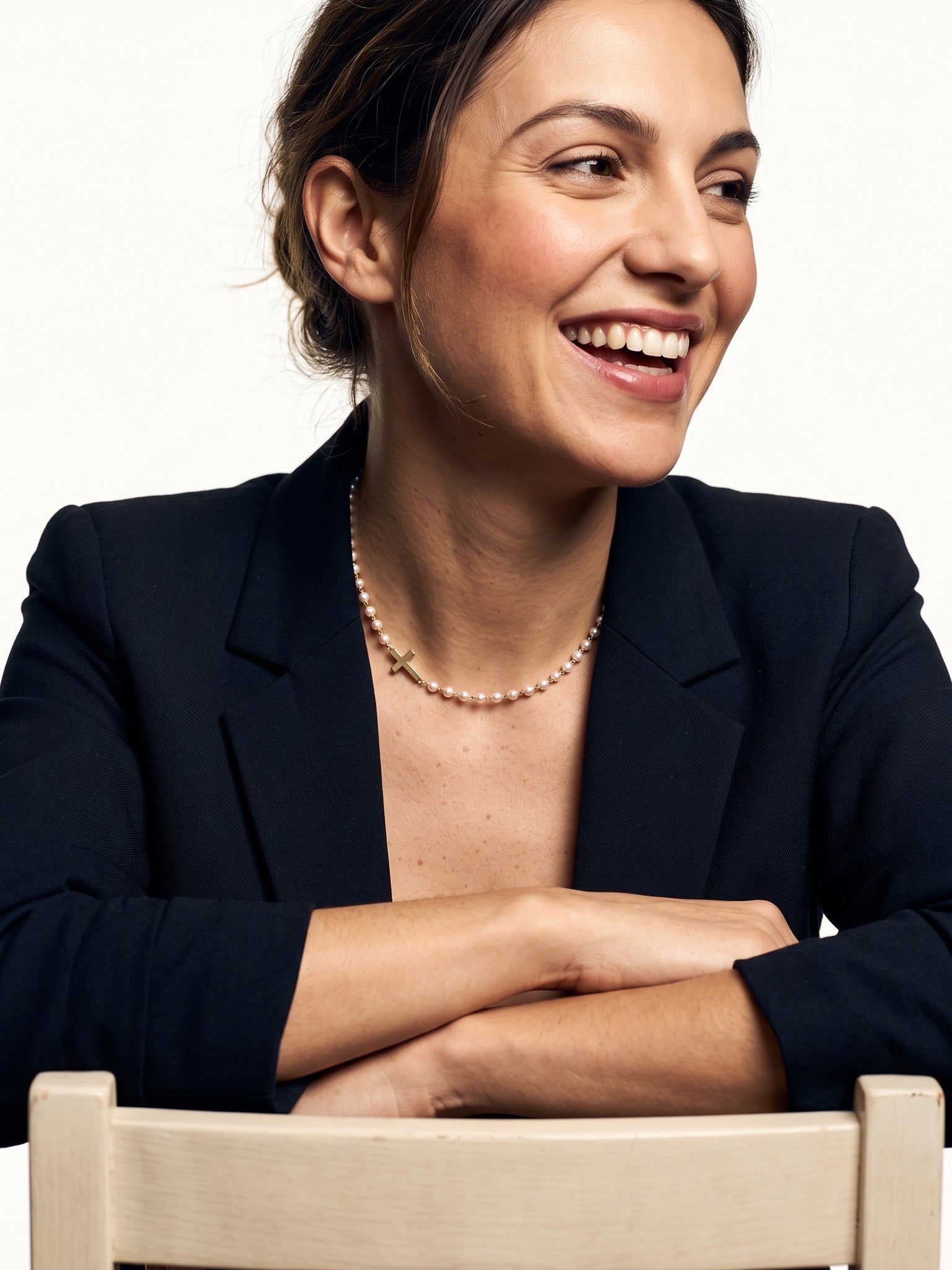 Every Season Cross Necklace – worn on the neck, laughing, arms folded on a chair back, gold, dark blazer, ivory studio background