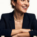 Every Season Cross Necklace – worn on the neck, laughing, arms folded on a chair back, gold, dark blazer, ivory studio background