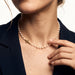Every Season Cross Necklace – worn at the collarbone, hand touching the pearls, gold, dark blazer, ivory studio background