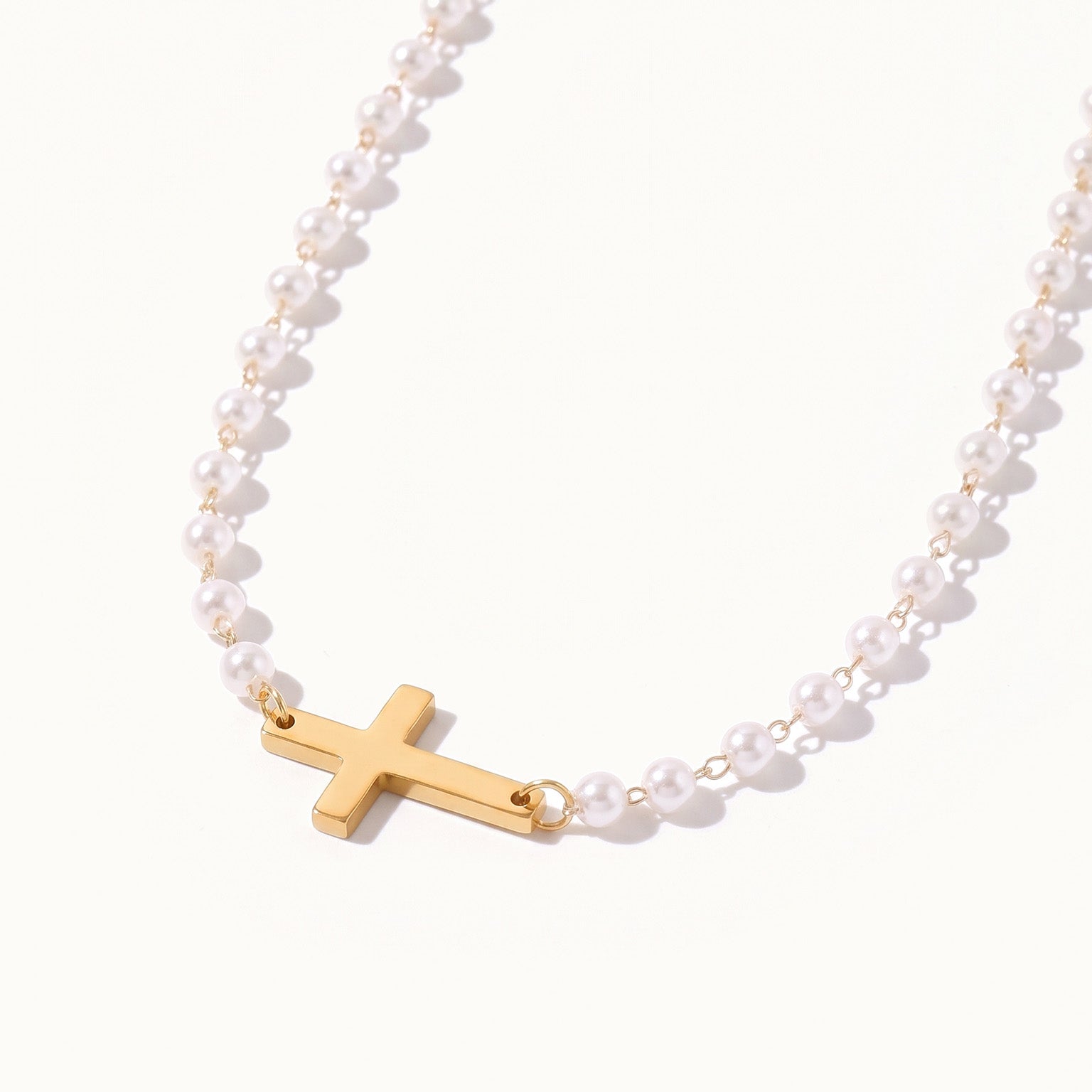Every Season Cross Necklace – gold cross on a strand of tiny pearls, hanging in a curve, on ivory background