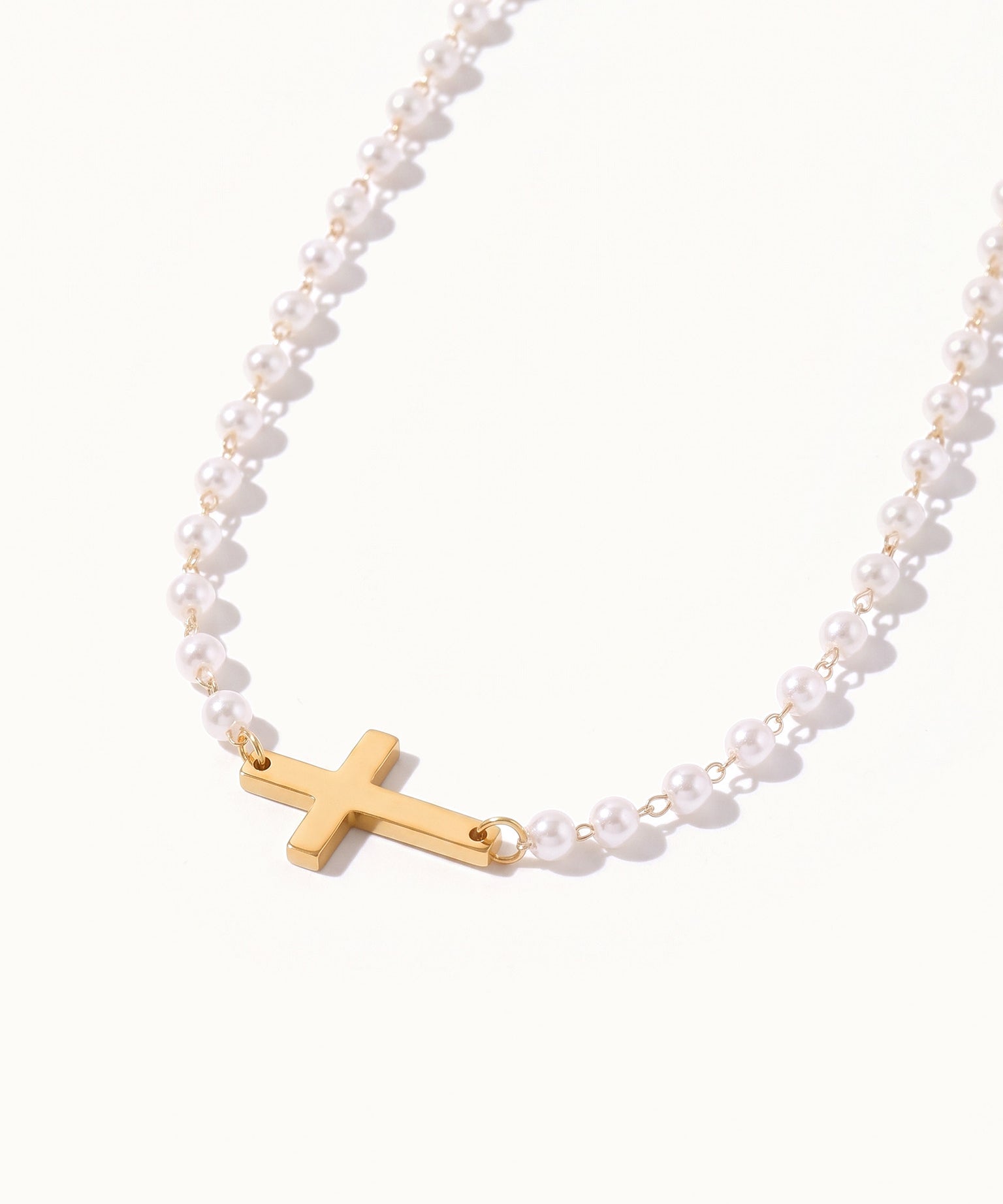 Every Season Cross Necklace – gold cross on a strand of tiny pearls, hanging in a curve, on ivory background