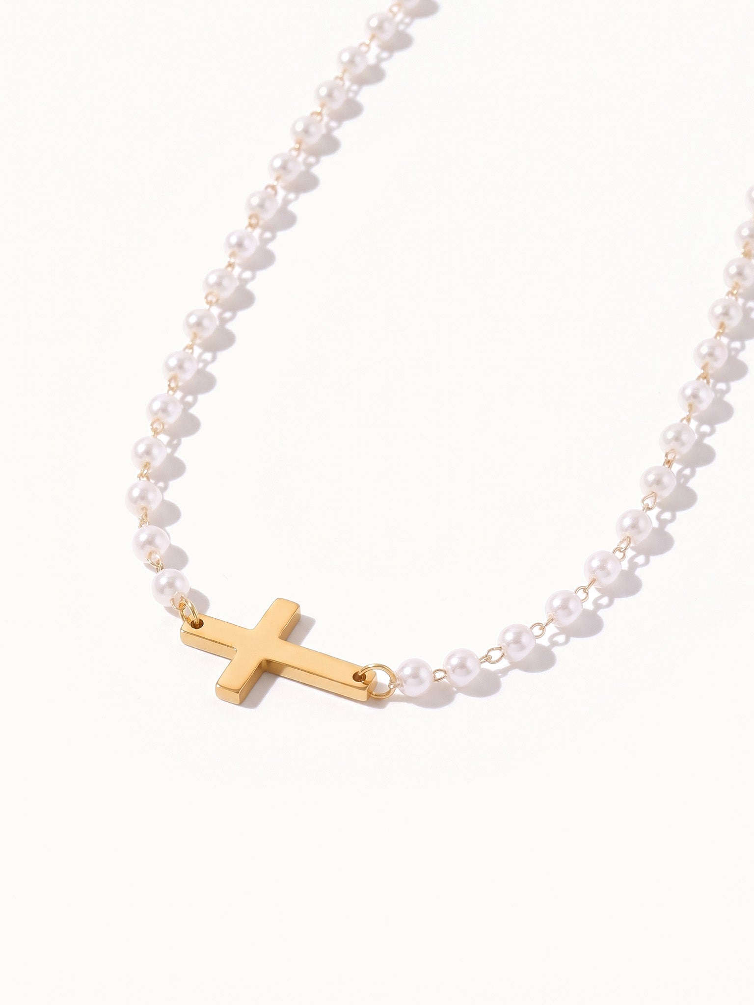 Every Season Cross Necklace – gold cross on a strand of tiny pearls, hanging in a curve, on ivory background