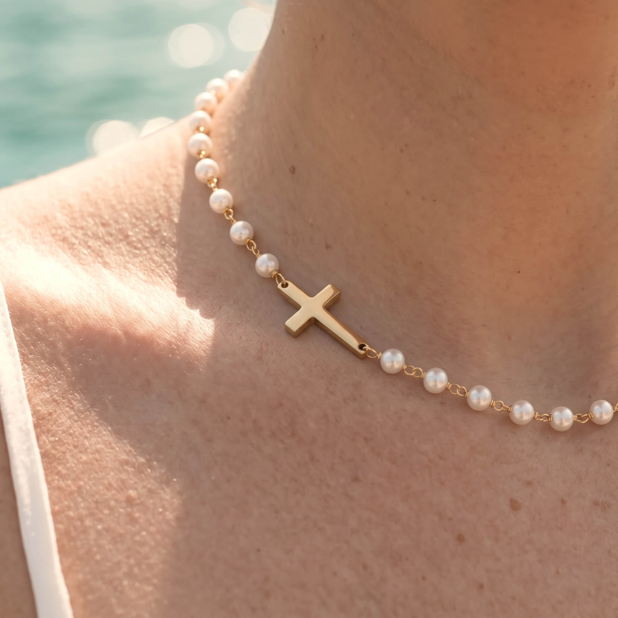 Every Season Cross Necklace – worn at the collarbone, gold cross and pearls, sunlit water behind