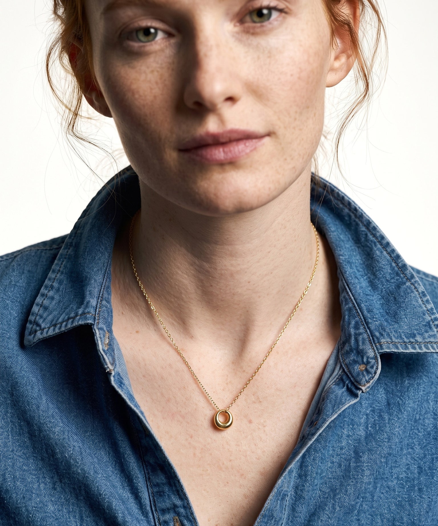 Everyday Loop Necklace – gold loop pendant on a fine chain at the collarbone, model with red hair in a blue denim shirt, ivory studio background