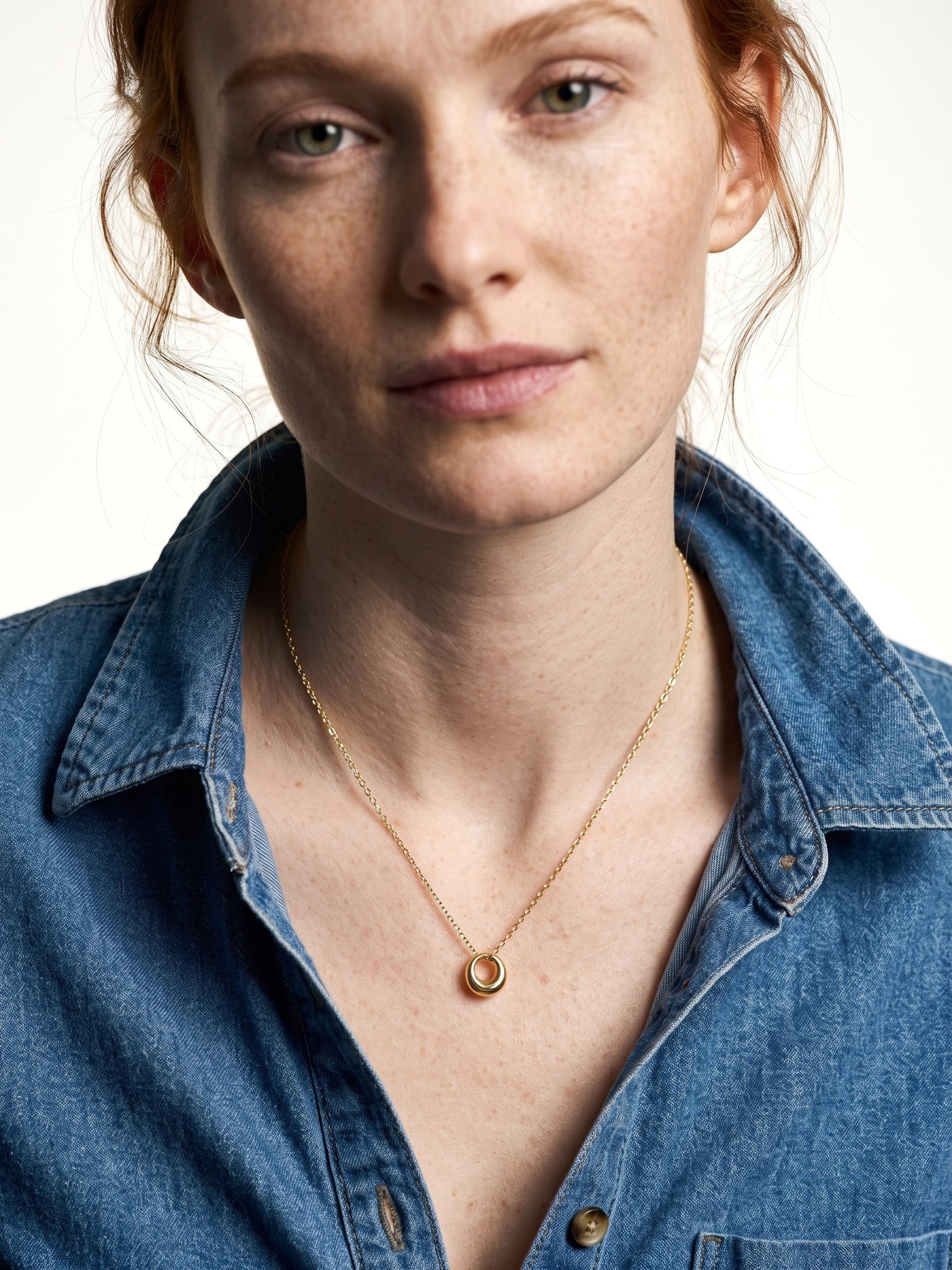 Everyday Loop Necklace – gold loop pendant on a fine chain at the collarbone, model with red hair in a blue denim shirt, ivory studio background