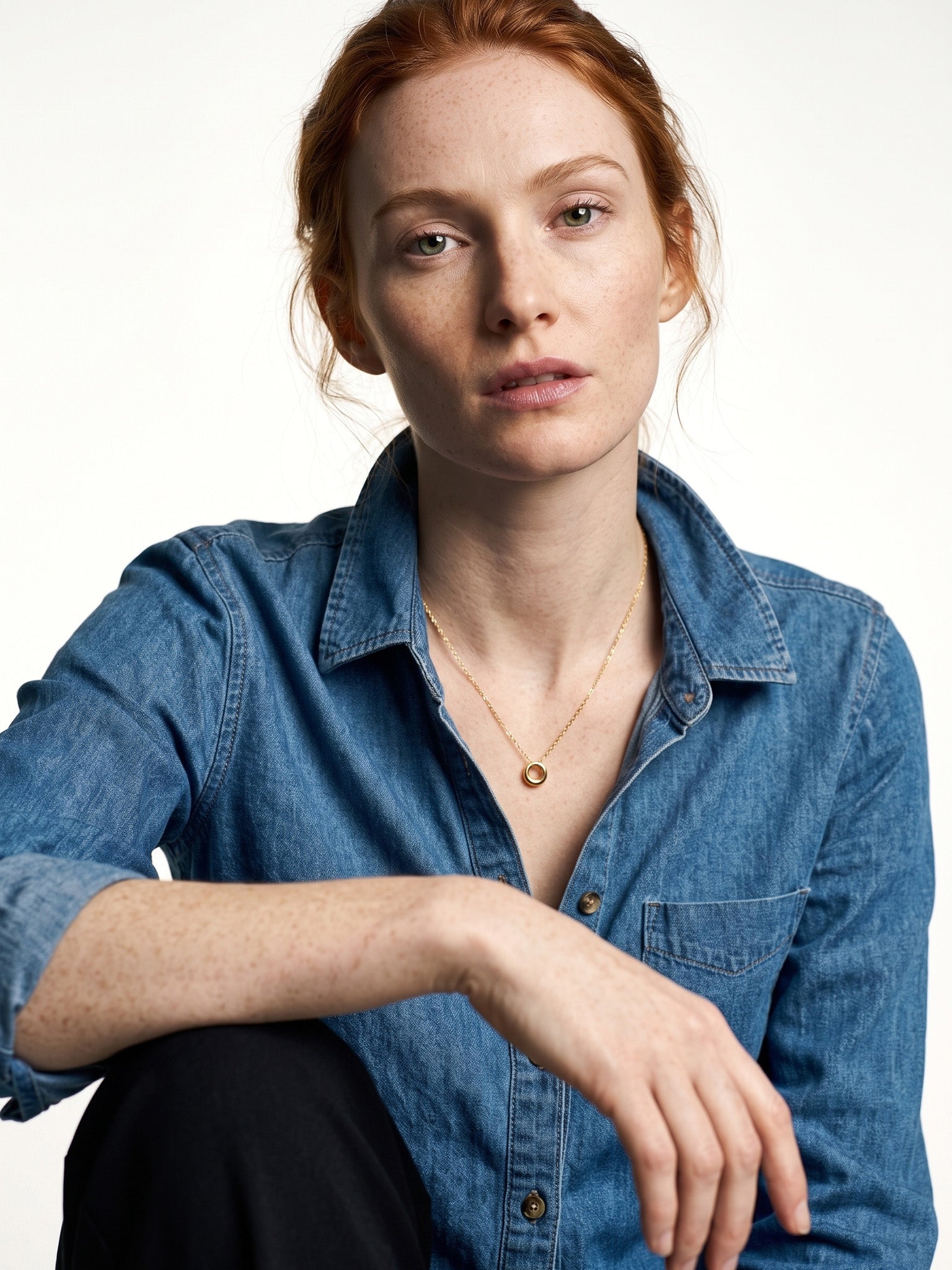 Everyday Loop Necklace – gold loop pendant on a fine chain, model seated with one arm resting on her knee, blue denim shirt, ivory studio background