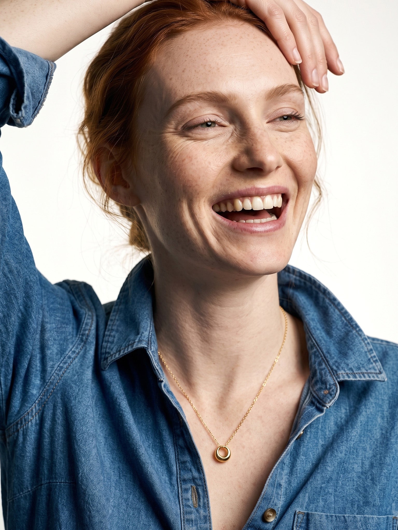Everyday Loop Necklace – gold loop pendant on a fine chain, laughing model with one hand resting on her head, blue denim shirt, ivory studio background