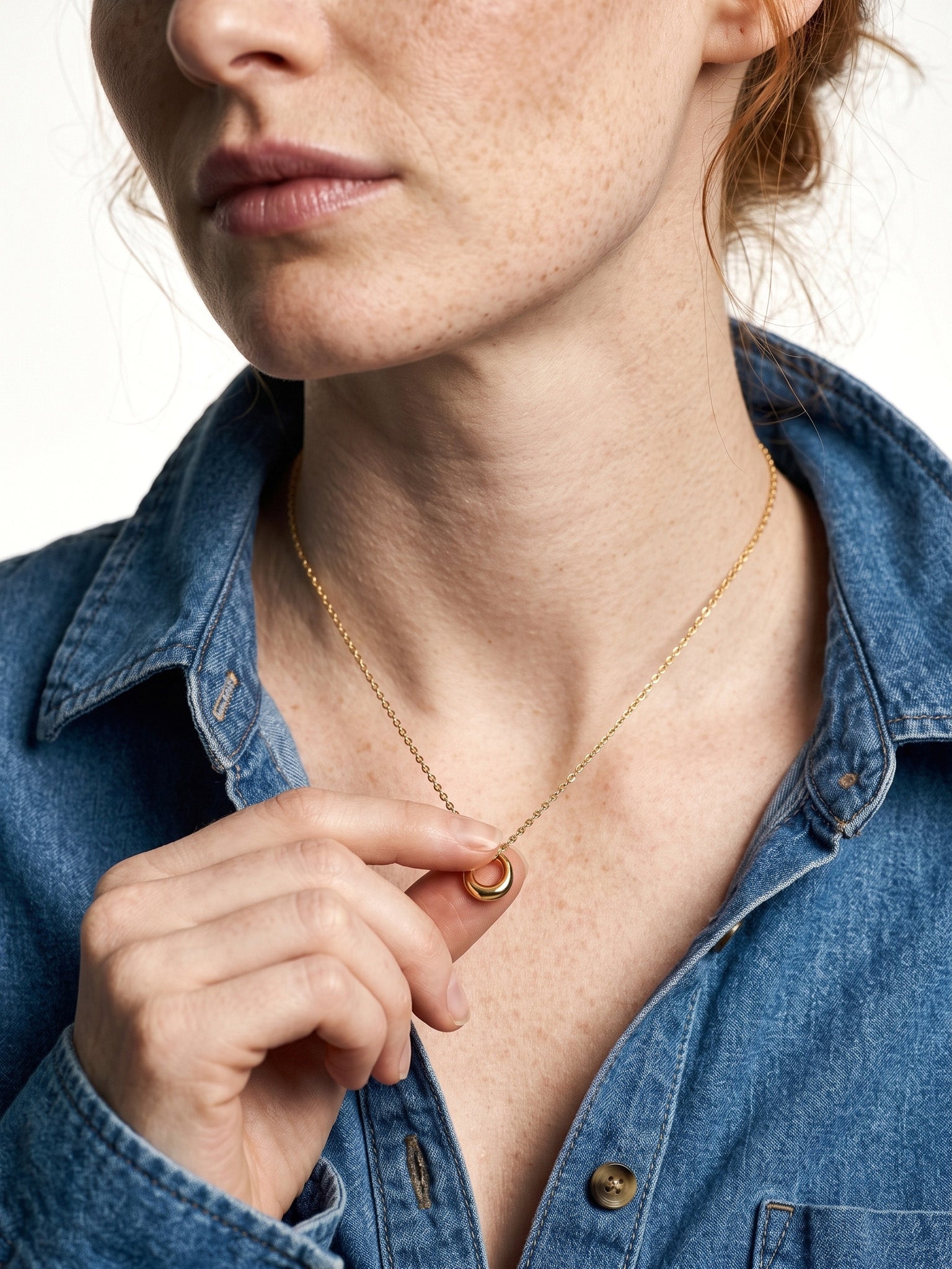 Everyday Loop Necklace – fingers holding the gold loop pendant on its fine chain, blue denim shirt, ivory studio background