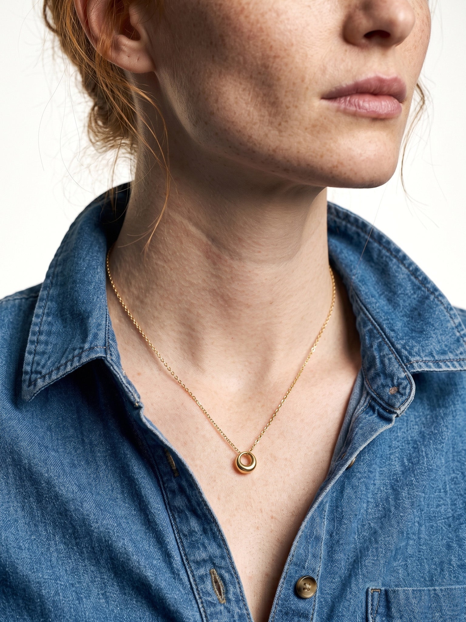 Everyday Loop Necklace – gold loop pendant on a fine chain, model with her face turned to one side, blue denim shirt, ivory studio background