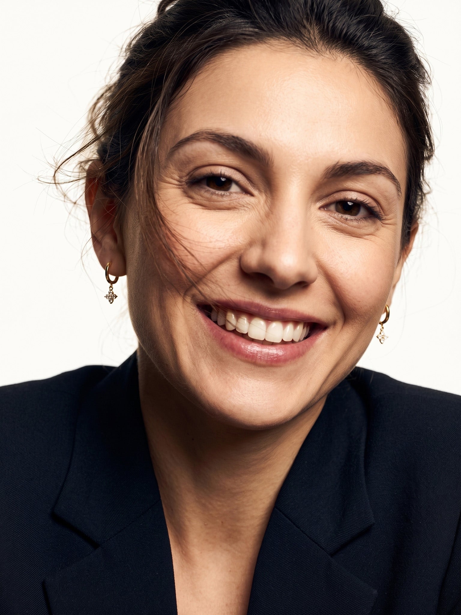 Field Lily Drop Earrings – worn in both ears, broad smile to the camera, gold huggies with four-stone flower drops, dark hair up