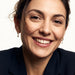 Field Lily Drop Earrings – worn in both ears, broad smile to the camera, gold huggies with four-stone flower drops, dark hair up