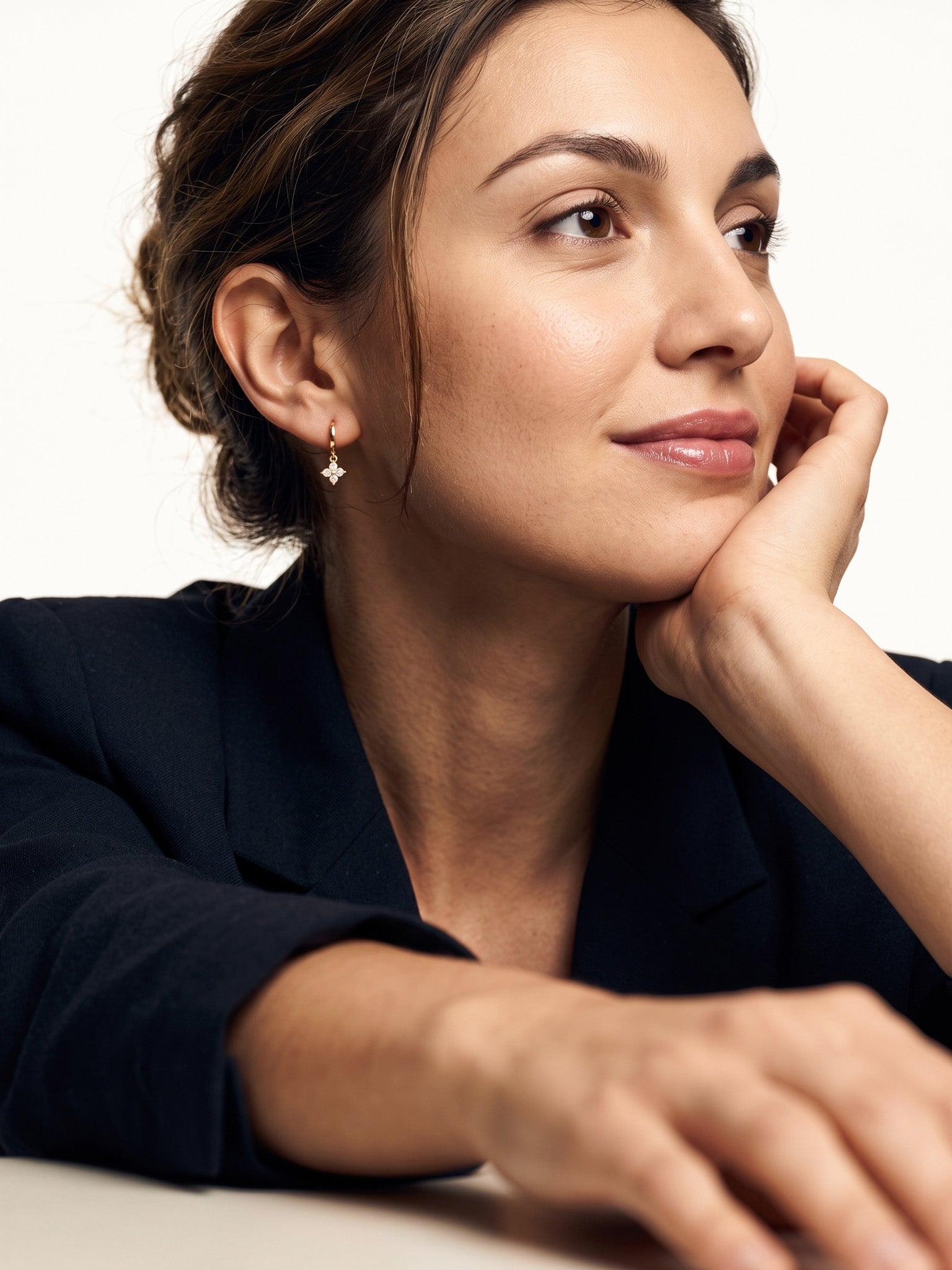 Field Lily Drop Earrings – worn in the ear, chin resting on the hand and looking away, gold huggie with a four-stone flower drop, dark hair up