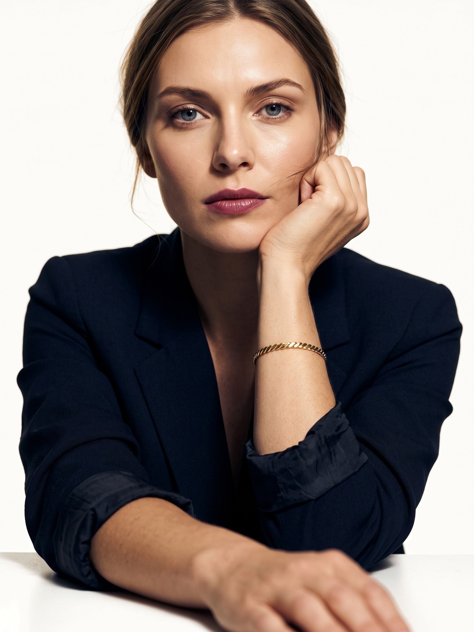 Fire & Roots Cuff – worn on the wrist, chin resting on the hand, seated at a table, gold, dark blazer, ivory studio background