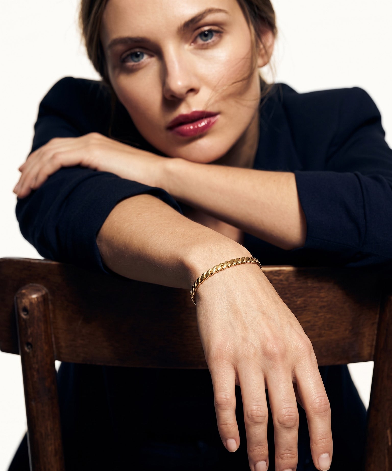 Fire & Roots Cuff – worn on the wrist, arm resting over the back of a wooden chair, gold, dark blazer, ivory studio background