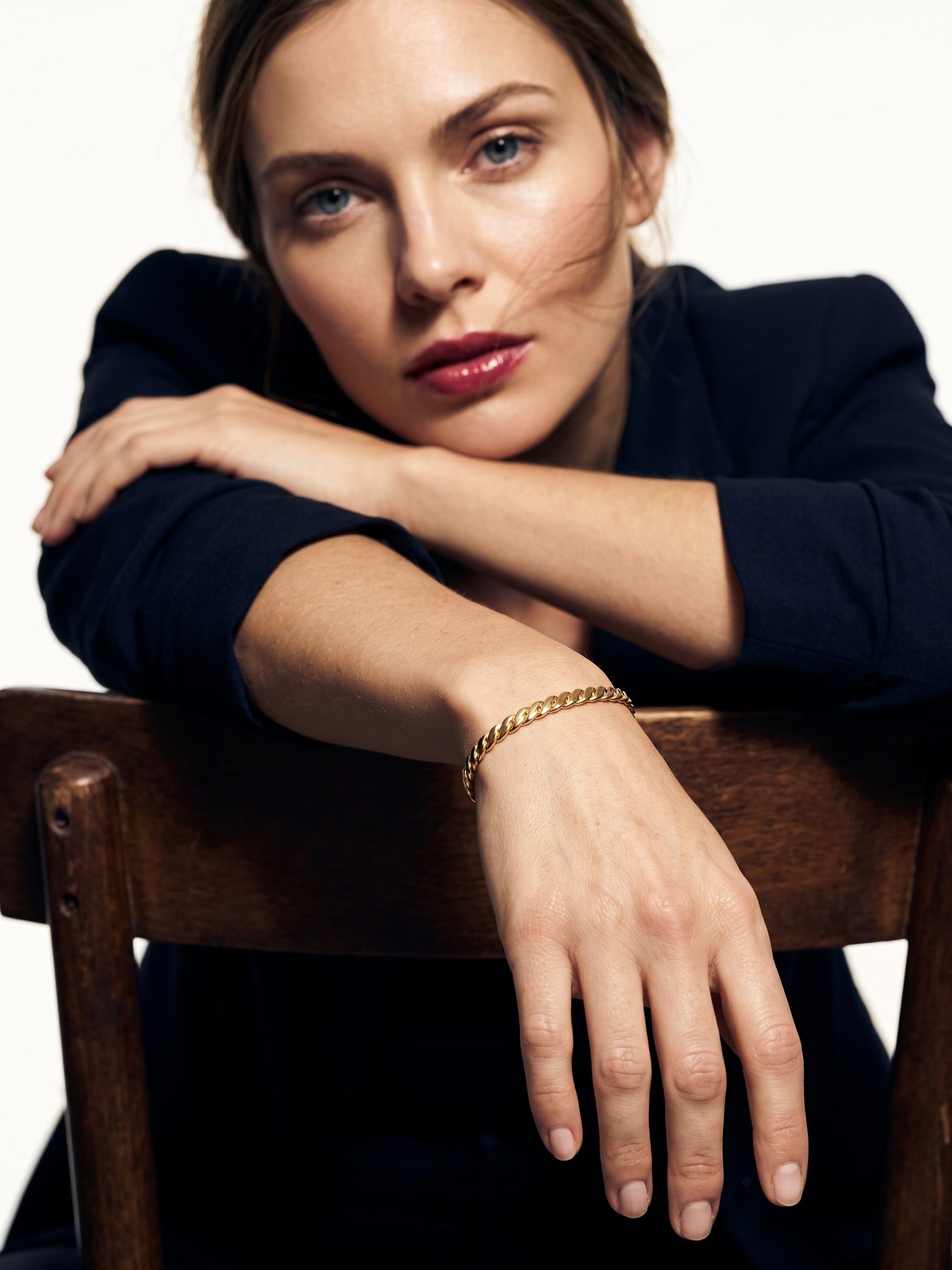 Fire & Roots Cuff – worn on the wrist, arm resting over the back of a wooden chair, gold, dark blazer, ivory studio background