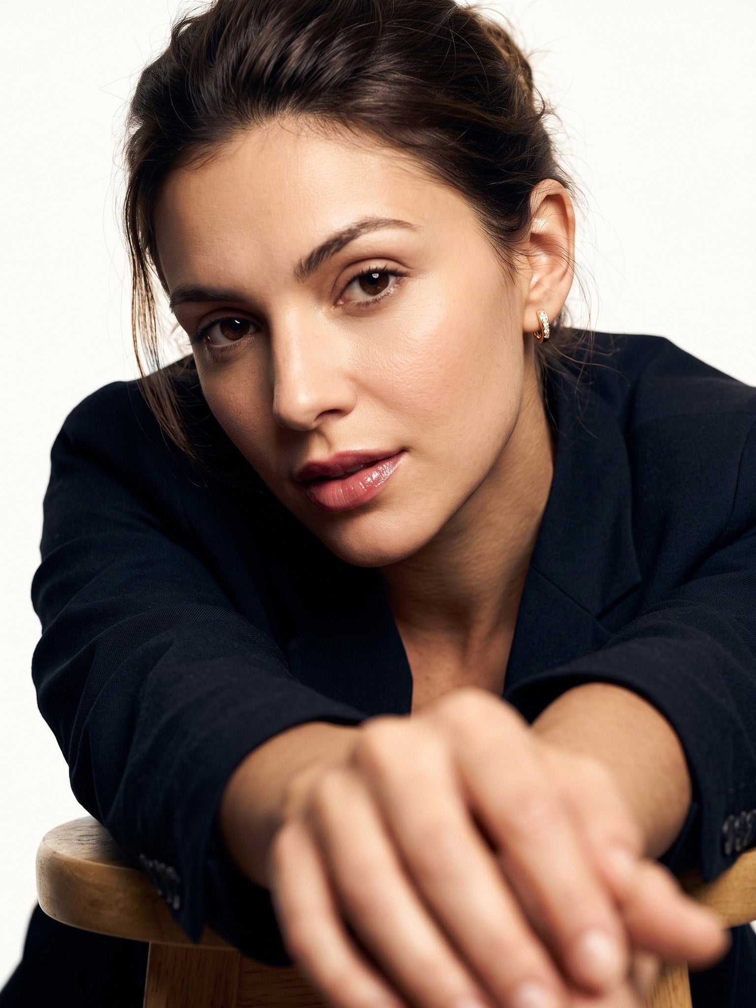 Four Seasons Huggies – worn in the ear, leaning forward on a wooden stool looking at the camera, gold four-stone huggie, dark hair up