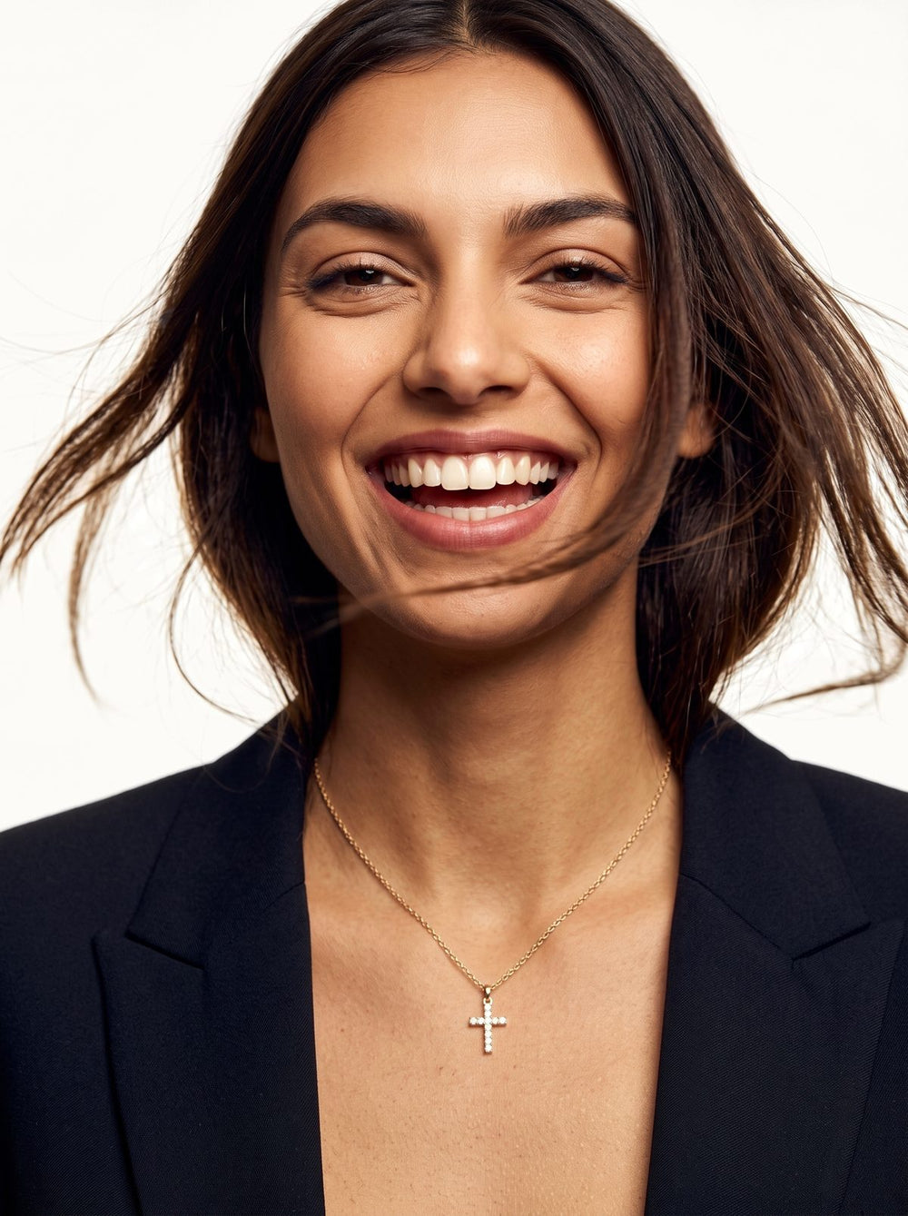 Woman laughing with her hair blowing, wearing a gold cross necklace and a black blazer