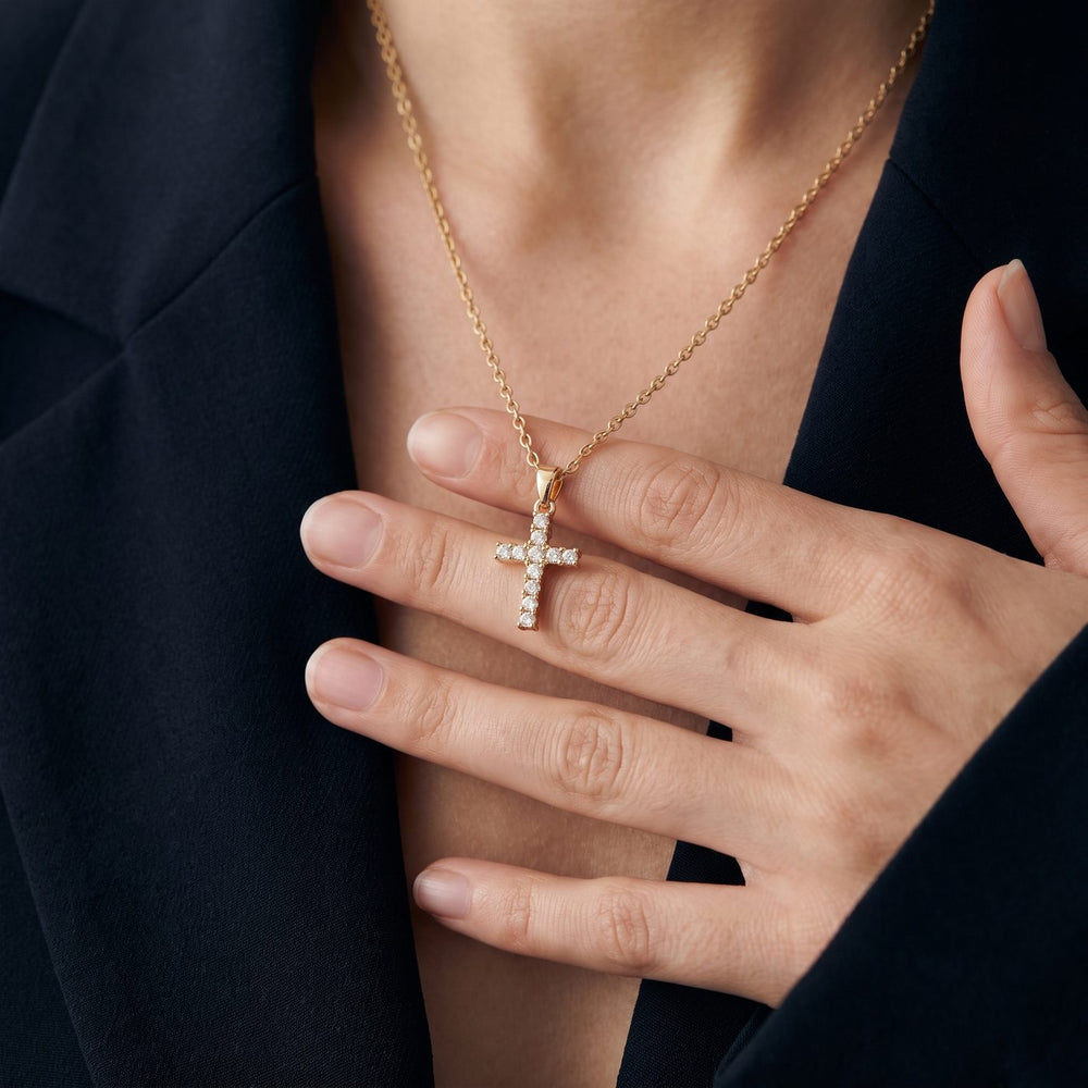 Hand holding a gold cross necklace with clear stones at the collarbone, black blazer