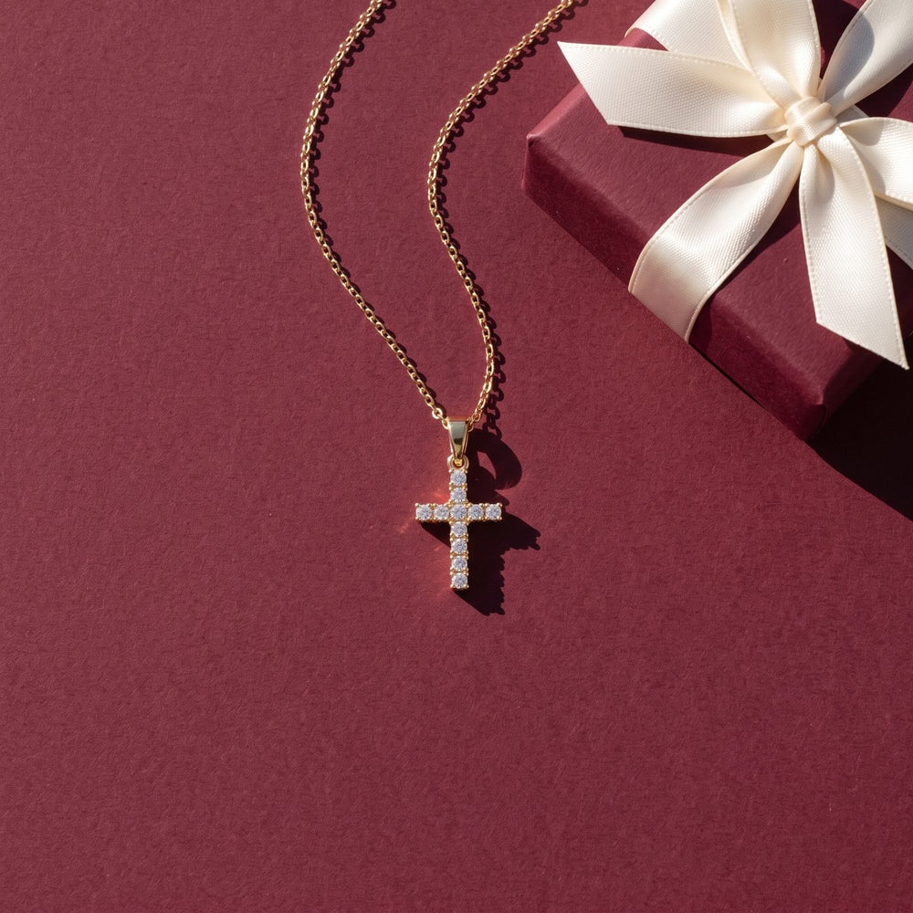 Gold cross necklace with clear stones on a burgundy background beside a gift box with an ivory ribbon