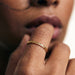 Golden Season Band Ring – worn with a finger raised to the lips, gold, close-up