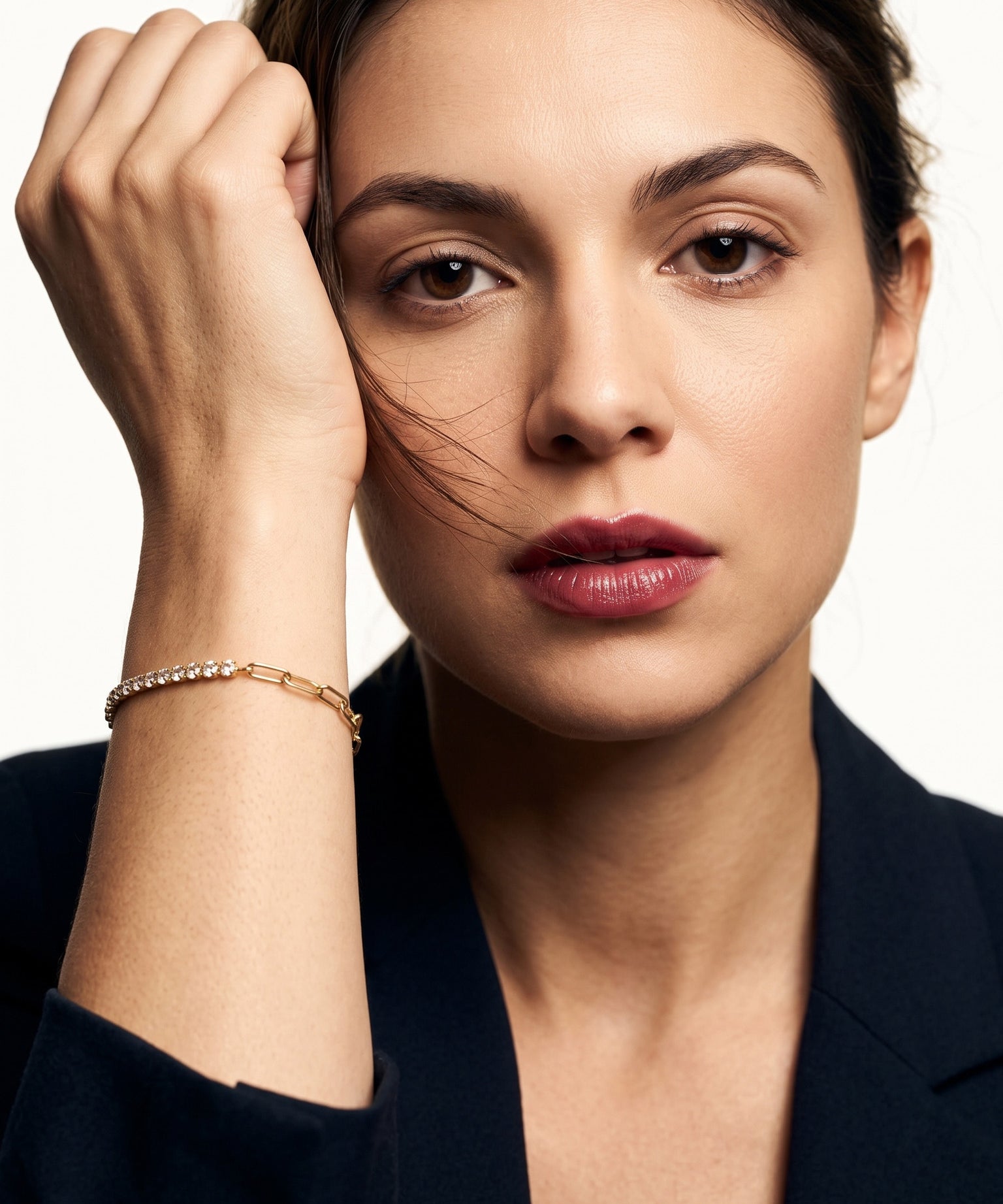 Grace Line Bracelet – worn on the wrist, hand raised to the forehead, gold, dark blazer, ivory studio background