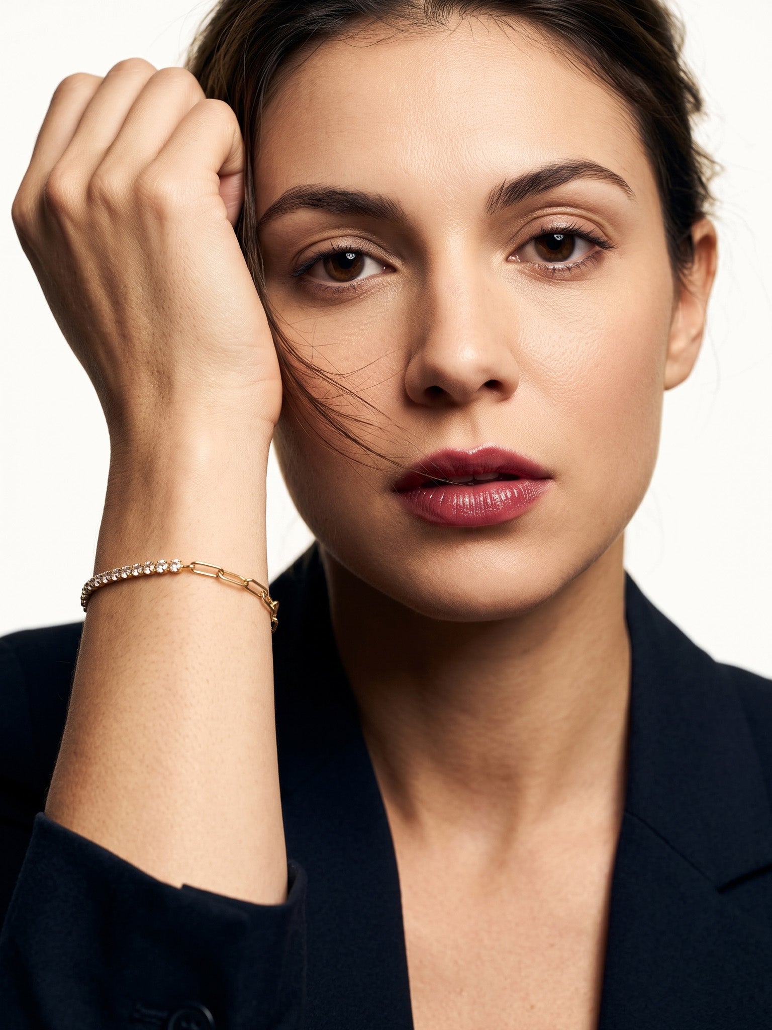 Grace Line Bracelet – worn on the wrist, hand raised to the forehead, gold, dark blazer, ivory studio background