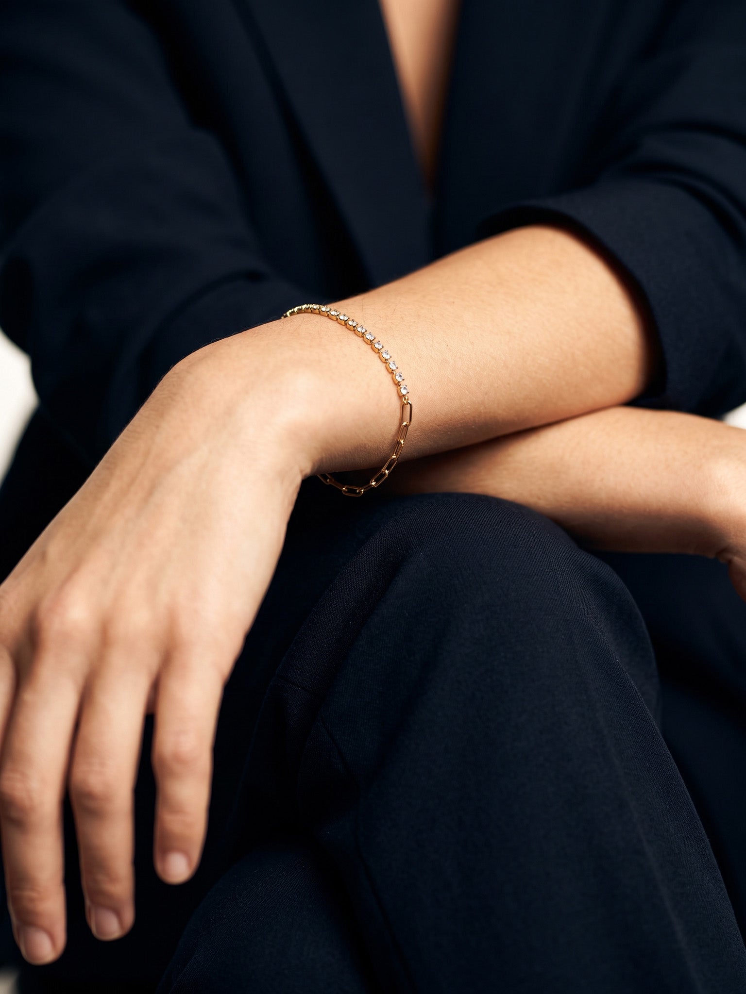 Grace Line Bracelet – worn on the wrist, arm resting across the knee, gold, dark blazer, ivory studio background