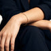 Grace Line Bracelet – worn on the wrist, arm resting across the knee, gold, dark blazer, ivory studio background