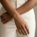 Grace Line Bracelet – worn on the wrist with arms crossed, gold, cream trousers