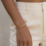 Grace Line Bracelet – worn on the wrist, hand relaxed at the side, gold, cream trousers