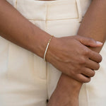Grace Line Bracelet – worn on the wrist, one hand holding the other arm, gold, cream trousers