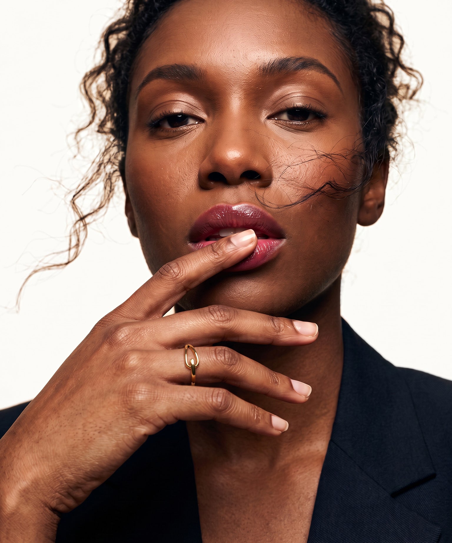 Held Ring – worn with a fingertip resting on the lips, gold, dark blazer, ivory studio background