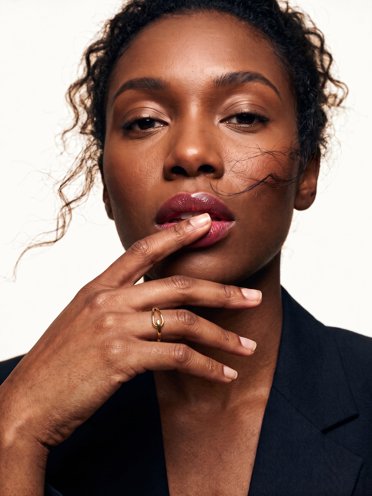 Held Ring – worn with a fingertip resting on the lips, gold, dark blazer, ivory studio background